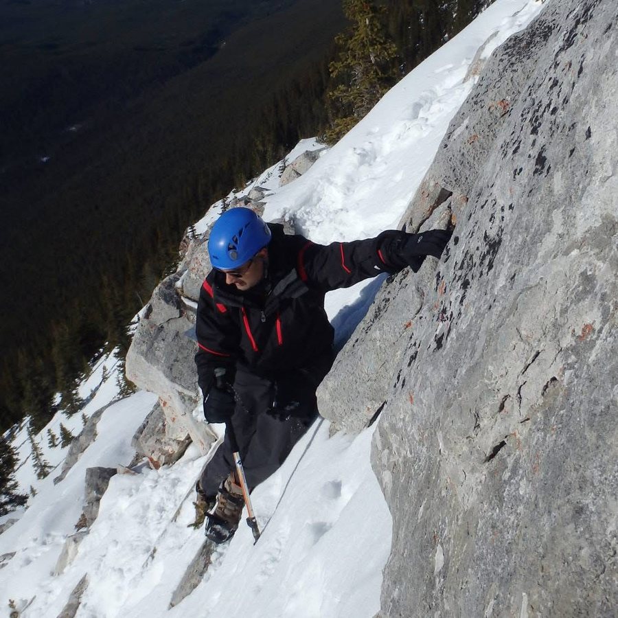 Scrambling up a snow-covered rock face in a helmet, with a valley and peaks below